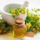 Small bottle of oil with cork lid, surrounded by st johns wort and a white mortar and pestle on a light wooden surface